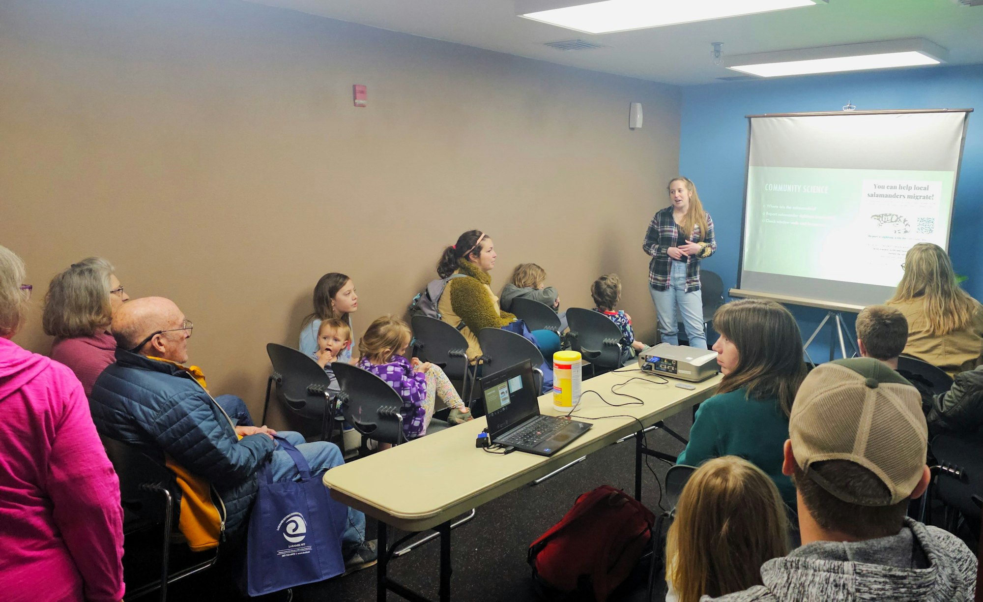 A presenter is addressing a mixed-age audience in a room with a projection about community science related to salamanders.