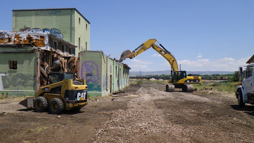 The image shows construction machinery, including an excavator and a skid loader, working to demolish a graffiti-covered building.