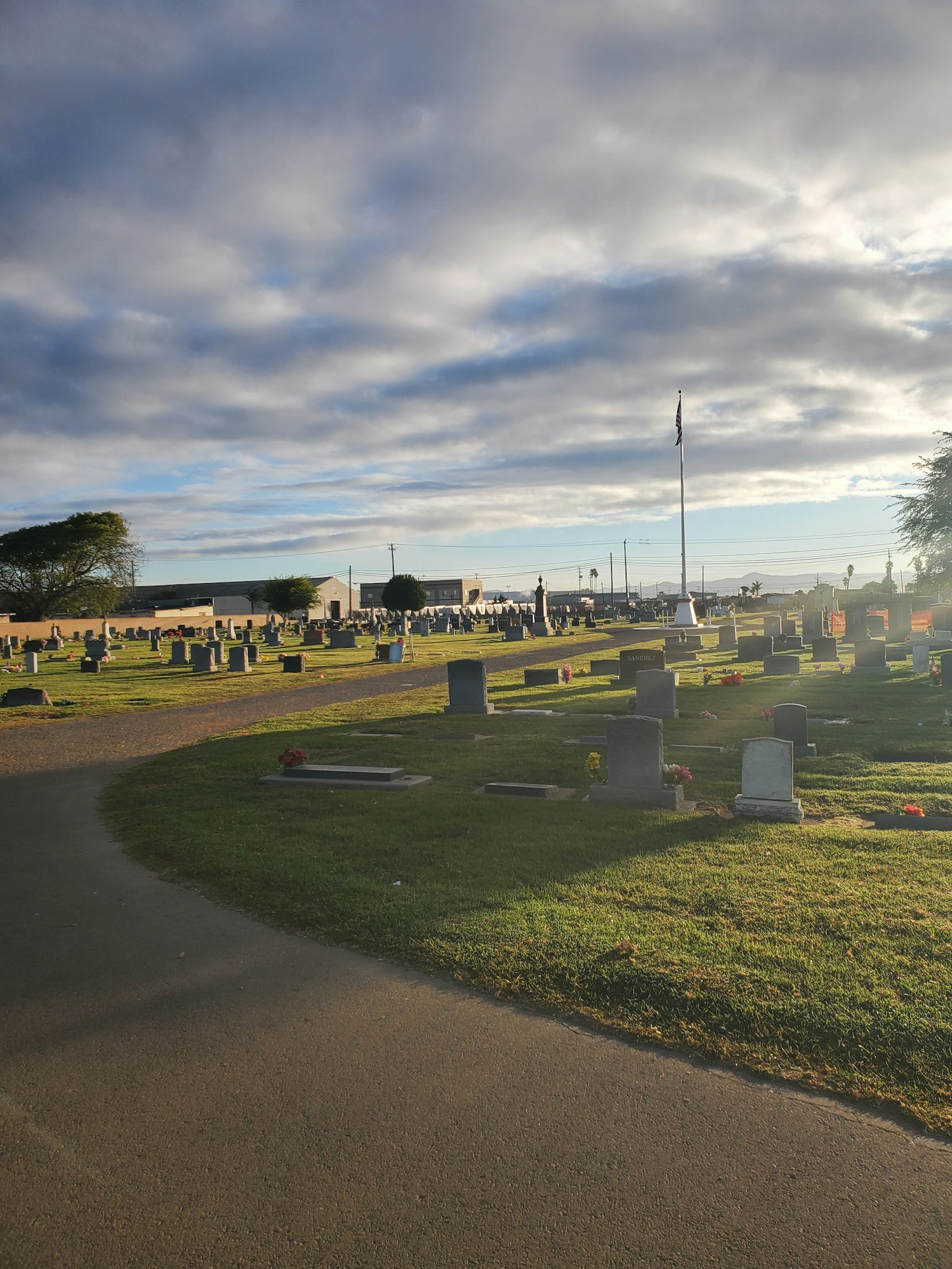 The image shows a cemetery with gravestones, surrounded by green grass and a cloudy sky in the background.