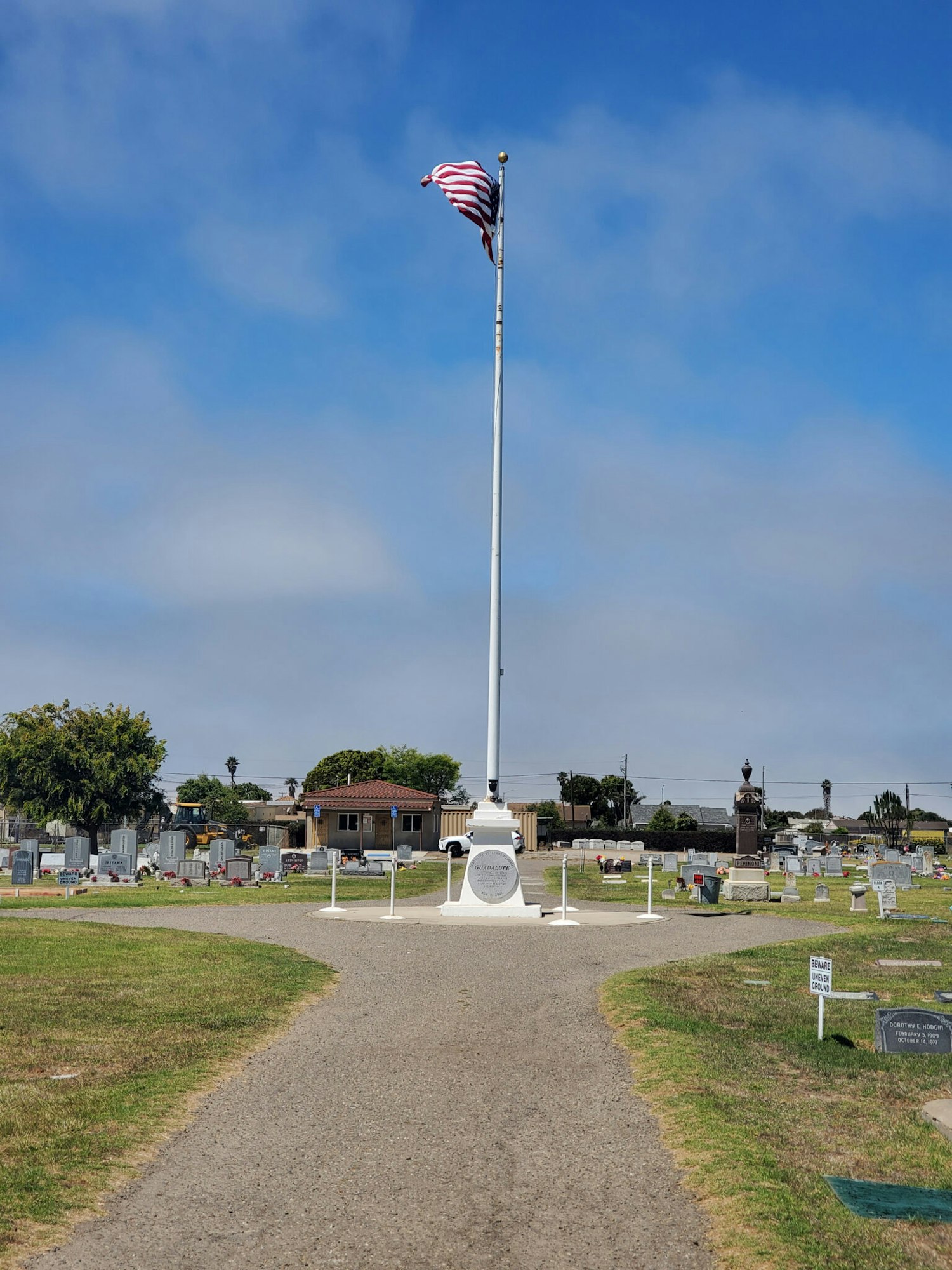 A flagpole with an American flag stands in a cemetery, surrounded by gravestones and a pathway leading through the grounds.