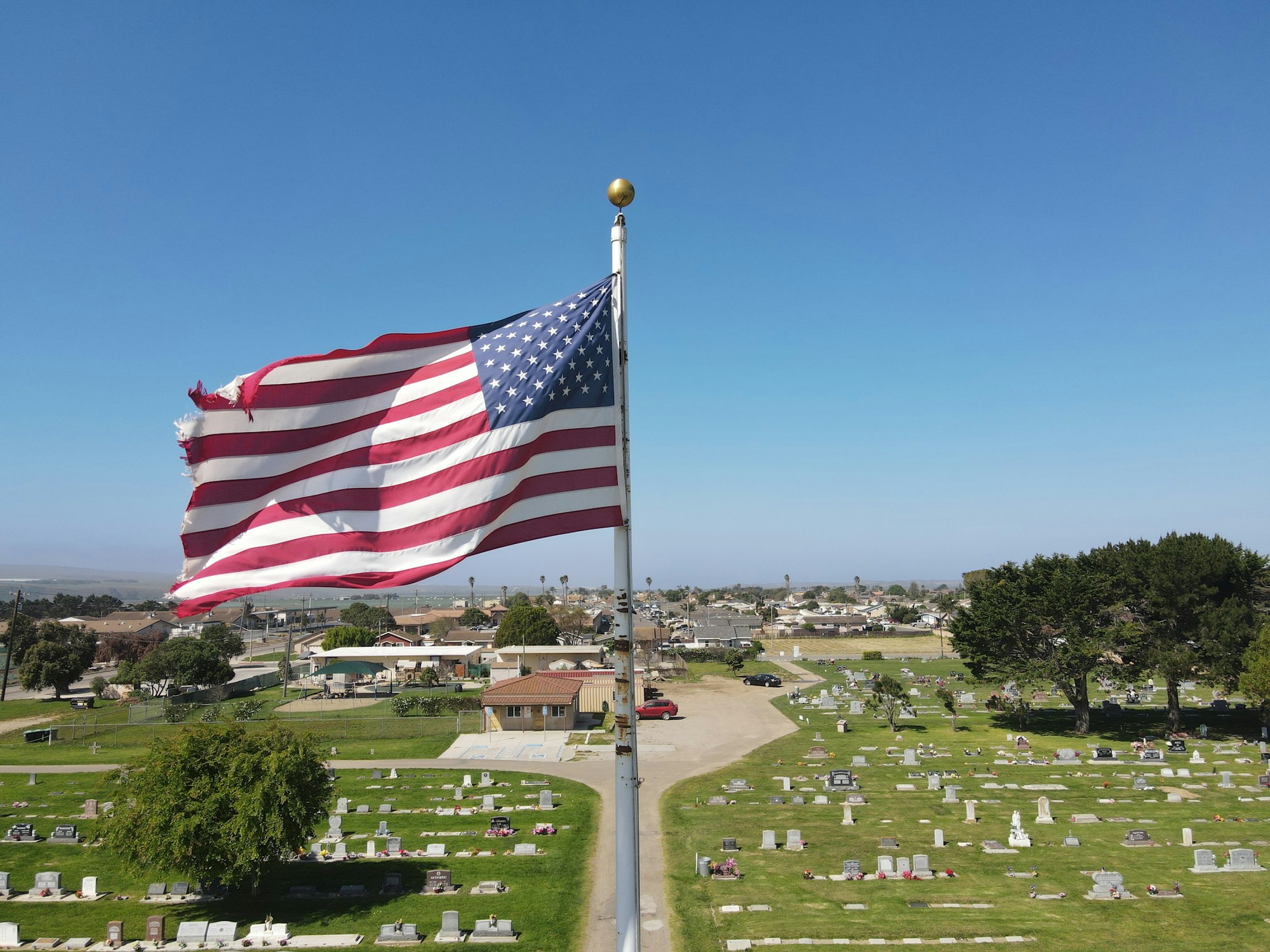 A tattered American flag flies over a cemetery, with a view of nearby buildings and a clear blue sky.