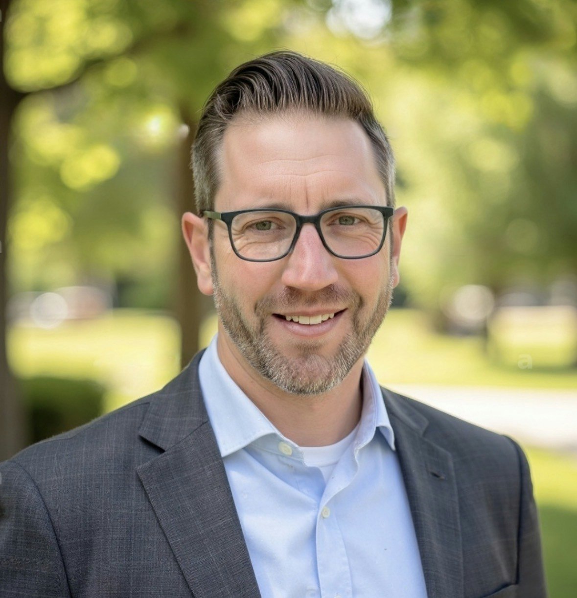 A smiling man with glasses, wearing a suit, outdoors with green trees and blurred background.