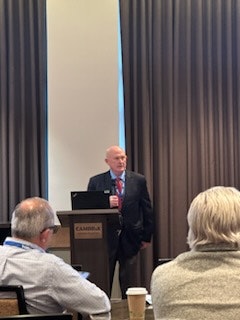 A person is giving a presentation at a lectern to an audience in a conference room setting.