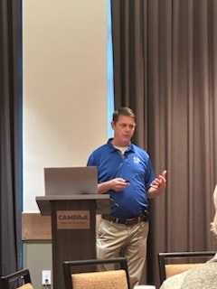 A person presenting at a podium in a room with chairs and a projector screen.