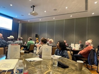 A group of people seated, facing forward, possibly in a conference room or seminar with papers on the tables.