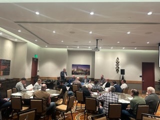 A meeting room with people seated around tables, listening to a presenter at the front.