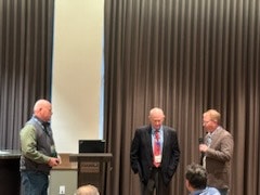 Three men on a stage; one standing at a podium, two others having a conversation. Curtain background, conference or event setting.