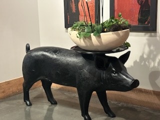 A sculpture of a pig with a bowl of plants on its back, inside a room with artwork on the wall.