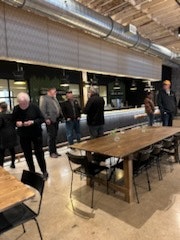 An indoor space with people standing by a counter, wooden tables in the foreground, and exposed ceiling ducts.