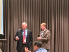 Two men speaking at a conference with an audience in the foreground.