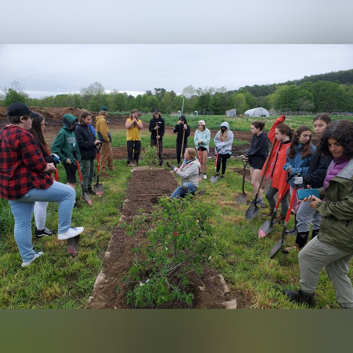 A group of people, mostly youths, gather in a garden with shovels, possibly learning about planting or gardening techniques.