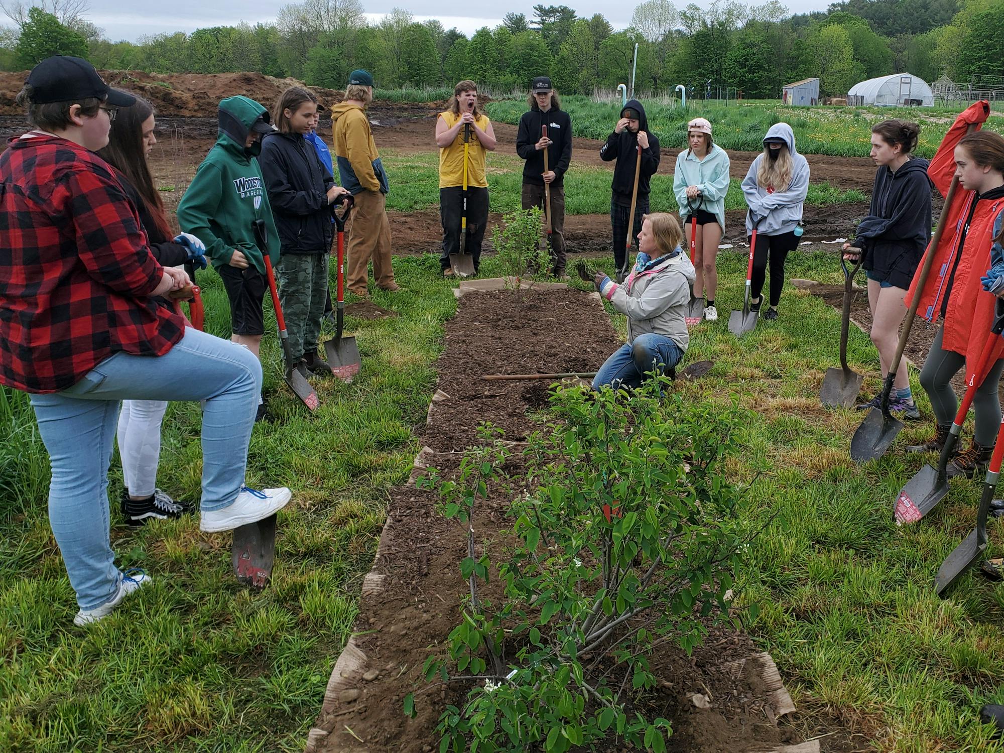 A group of people, mostly youths, gather in a garden with shovels, possibly learning about planting or gardening techniques.