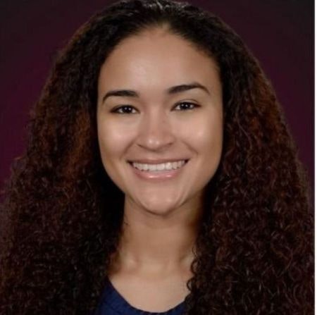 The image shows a smiling person with long, curly hair, wearing a dark top against a dark background.