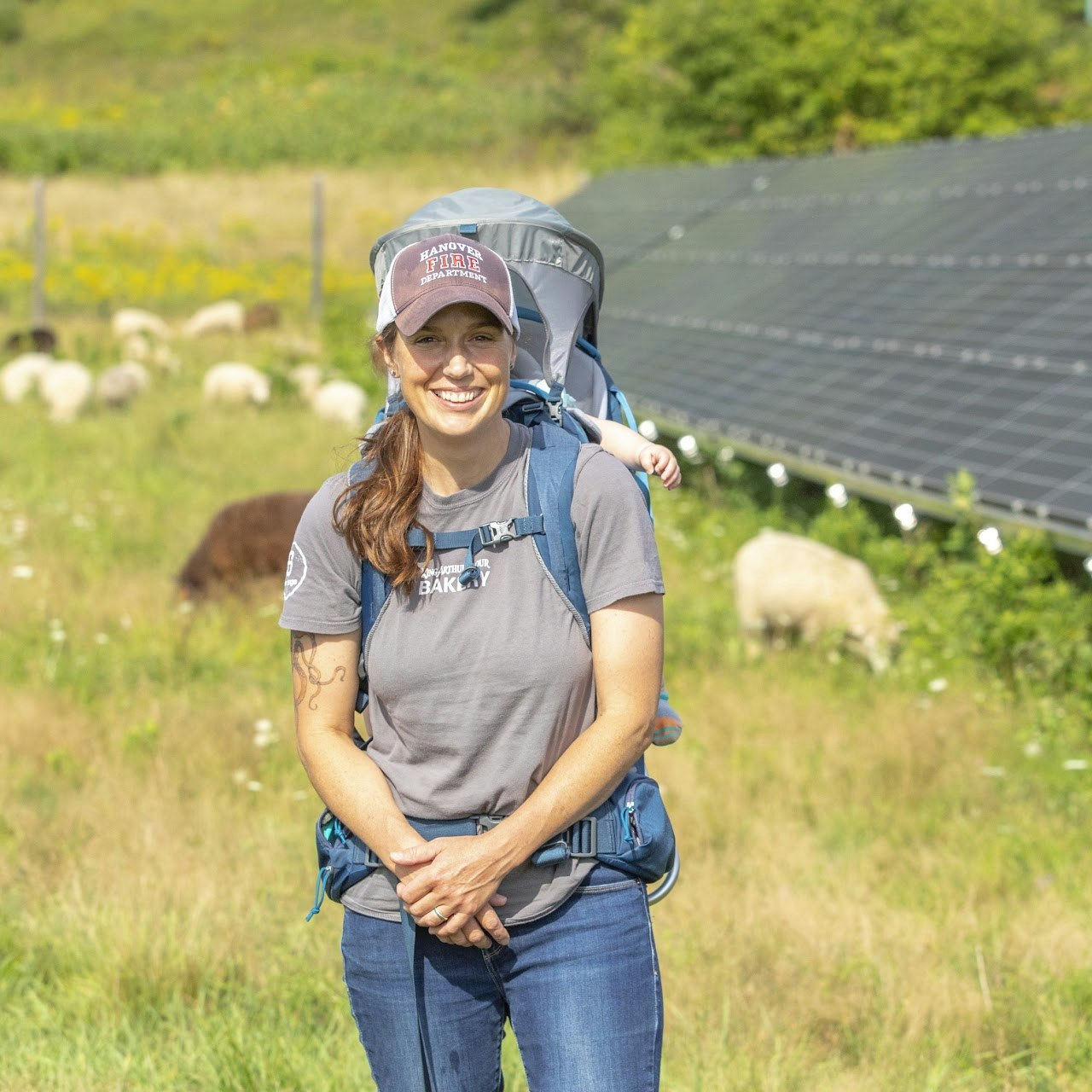A smiling woman stands in a field with sheep and solar panels, carrying a child in a backpack. It's a bright, sunny day.