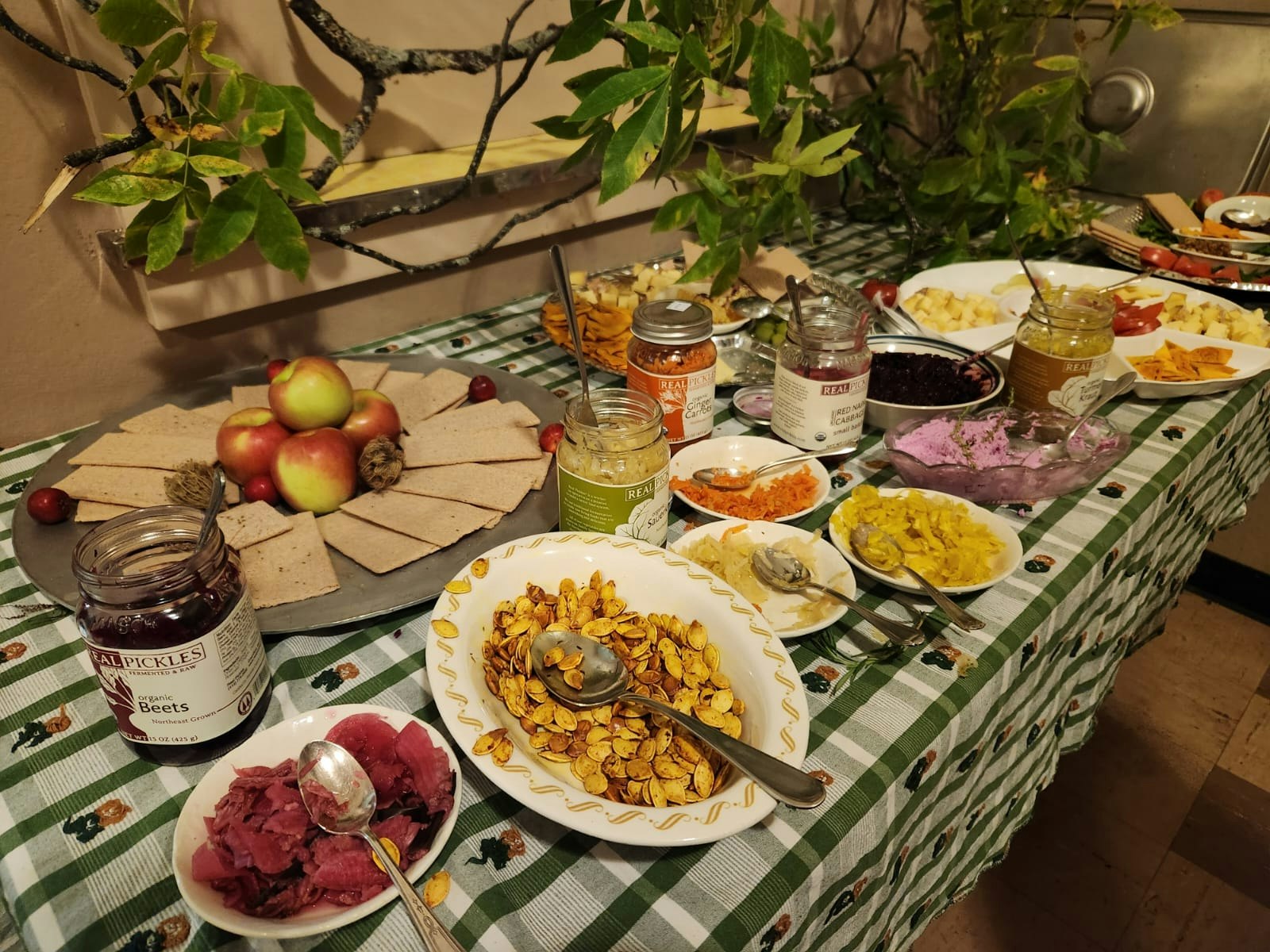 A table with jars of pickles, apples, crackers, and various spreads on a green checkered cloth.