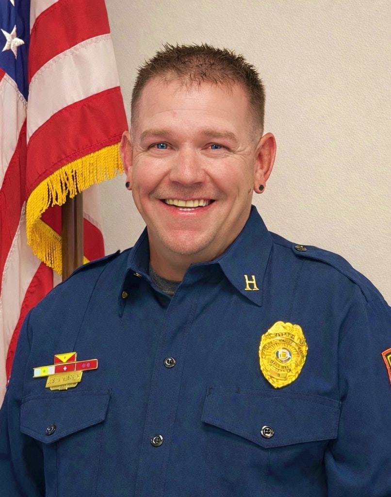 A smiling person in a blue uniform stands in front of an American flag, wearing badges and details on their uniform.