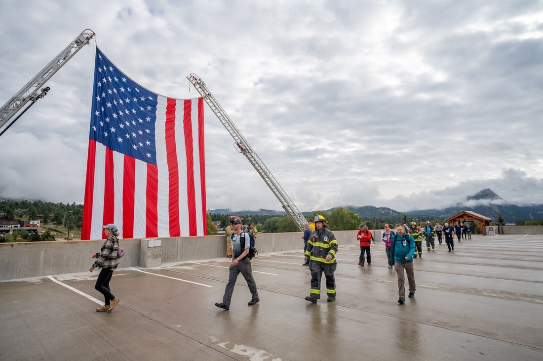 People walking under a large American flag hoisted by two cranes on a cloudy day.
