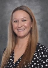 A person with long hair smiling, wearing a patterned top against a gray background.