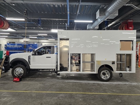 A large utility truck with a white boxy body, visible storage compartments, and a partially open side, inside a workshop.