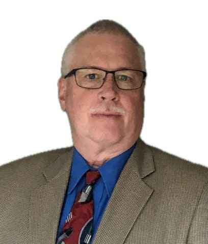 A person wearing glasses, a beige suit, blue shirt, and patterned tie, posing against a white background.