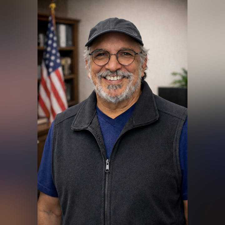 A smiling man with glasses and a cap, wearing a vest, stands in front of a bookshelf and an American flag.