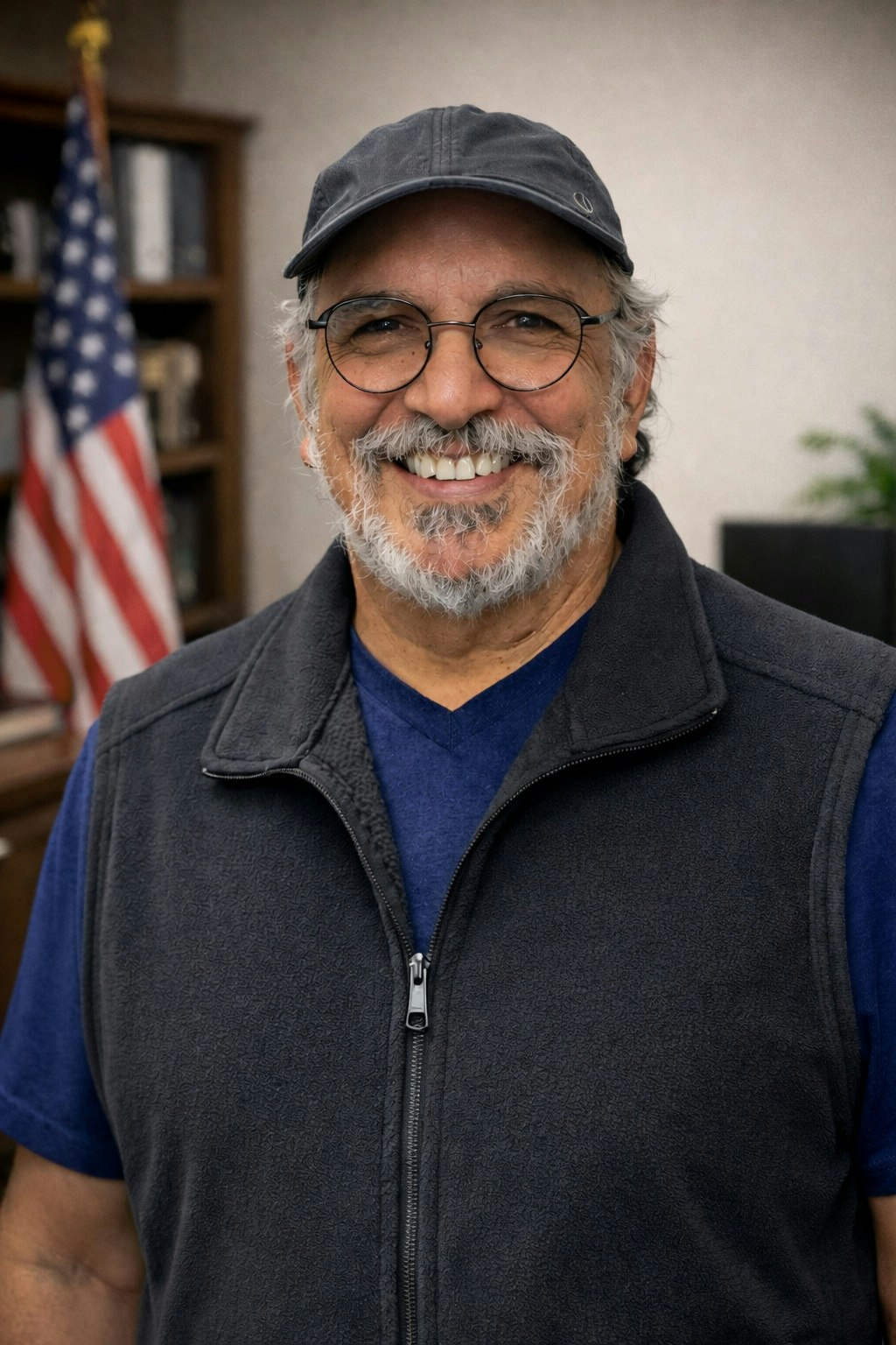 A smiling man with glasses and a cap, wearing a vest, stands in front of a bookshelf and an American flag.