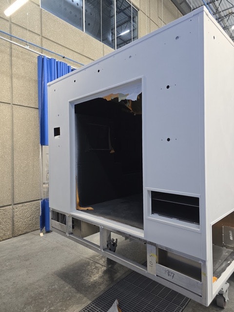 The image shows a large, mostly white structure with an open front, possibly a kiosk or display case, in a workshop or factory setting.