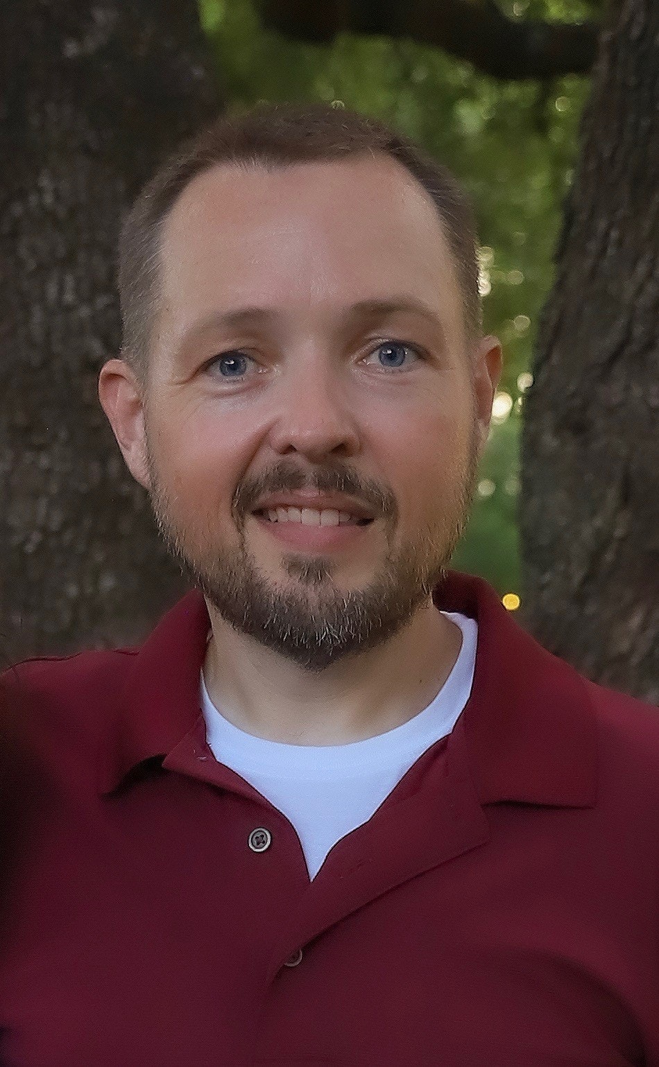 The image shows a man with a light beard, wearing a maroon polo shirt, smiling in front of blurred greenery.