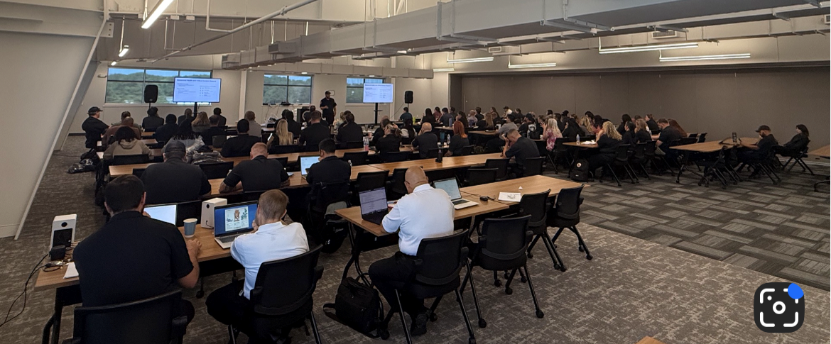 A large group of people sitting in a seminar room, focused on presentations, with laptops and notes in front of them.
