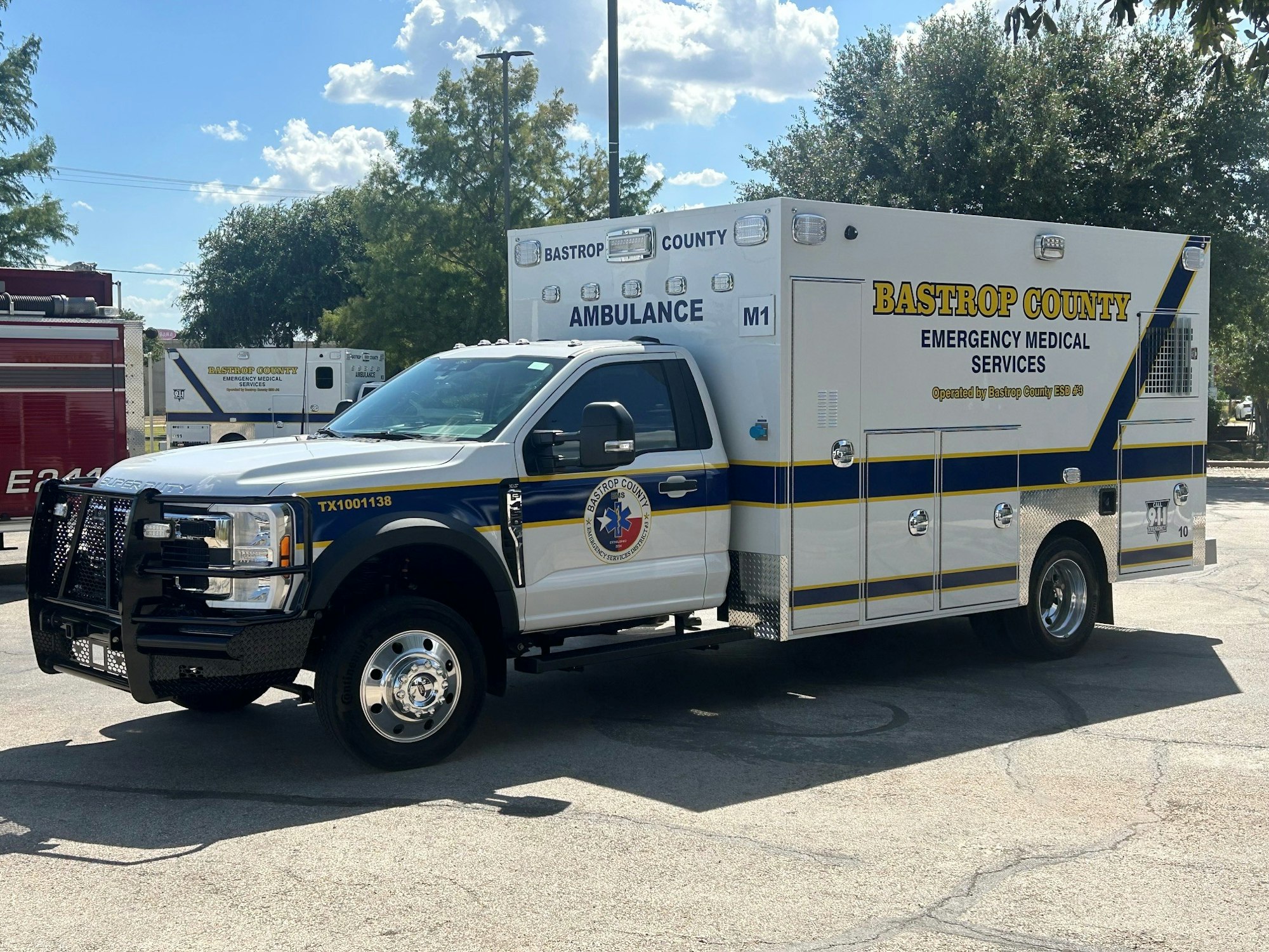 A Bastrop County ambulance vehicle designed for emergency medical services, parked in a lot with trees and other emergency vehicles nearby.