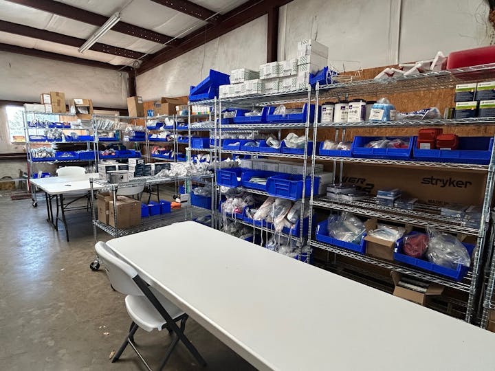 The image depicts a storage area with shelves filled with medical supplies and two tables in a well-organized space.