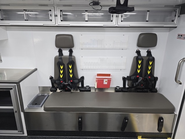 The image shows the interior of an ambulance with two seating areas equipped with safety harnesses and storage compartments.