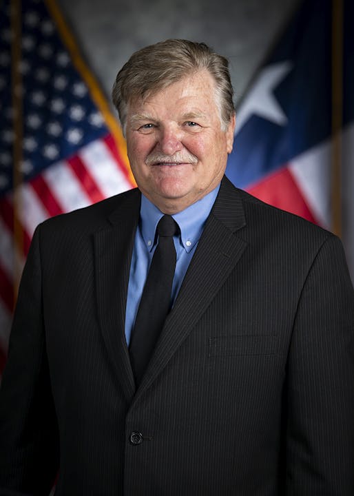 A man in a suit and tie stands in front of U.S. and Texas flags.