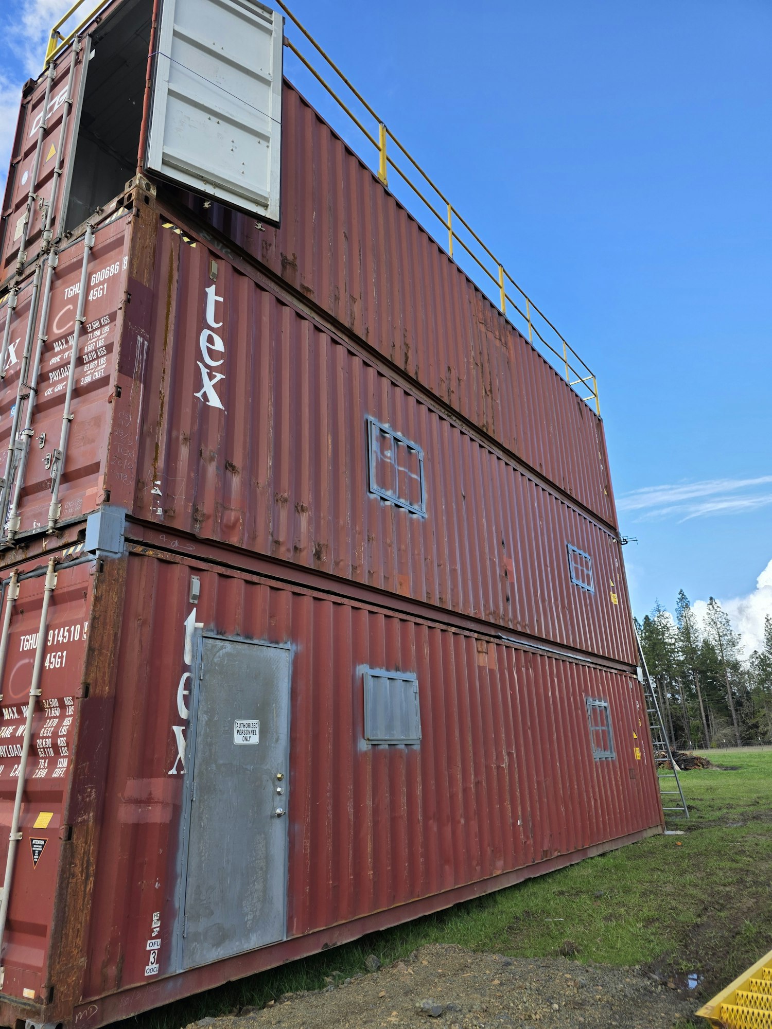 Stacked shipping containers with a door and window cutouts, set vertically in a grassy area under a blue sky.
