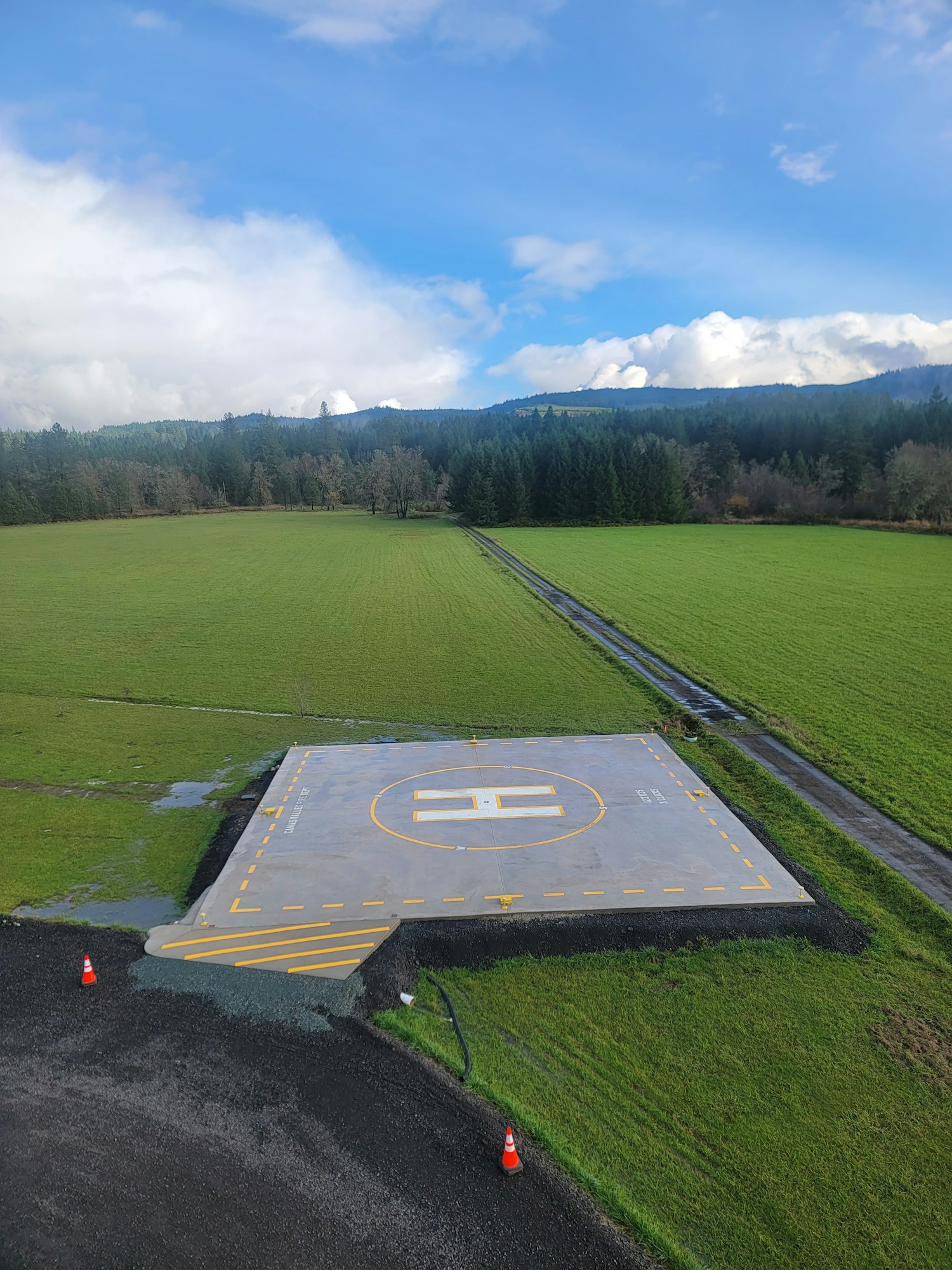 Concrete helipad in a grassy field, surrounded by traffic cones with trees and a cloudy blue sky in the background.
