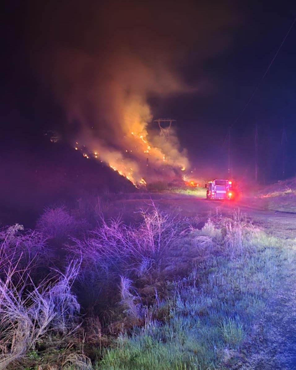 A wildfire at night with smoke, flames, and an emergency vehicle nearby.