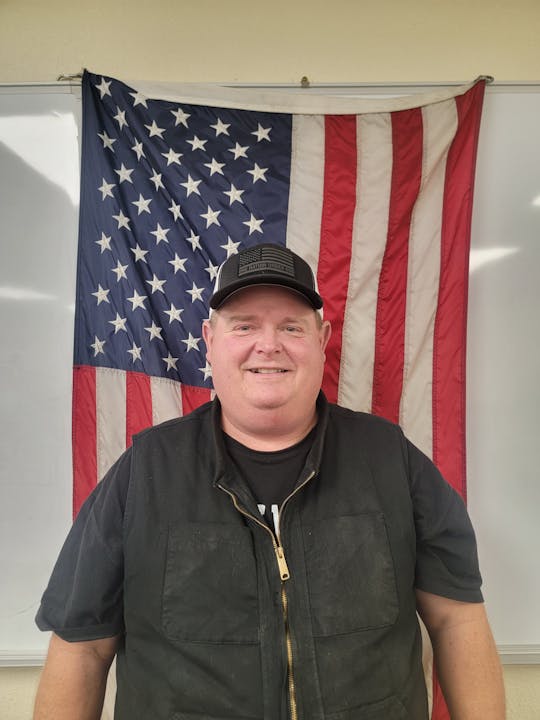 A person in front of an American flag, wearing a dark shirt and cap, smiles at the camera.