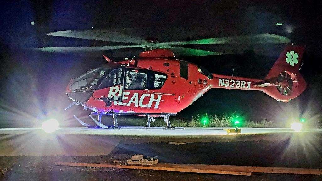 A red helicopter labeled "REACH" sits on a helipad at night, illuminated by green lights.