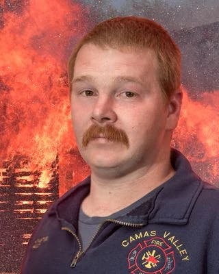 A person in a Camas Valley Fire uniform with a blazing fire in the background.