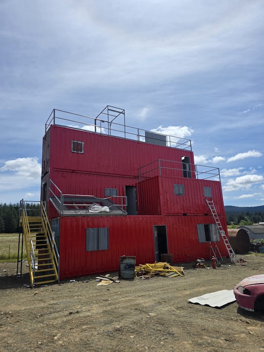 A red multi-story structure made of containers, with ladders and scaffolding, set on a dirt ground under a blue sky.