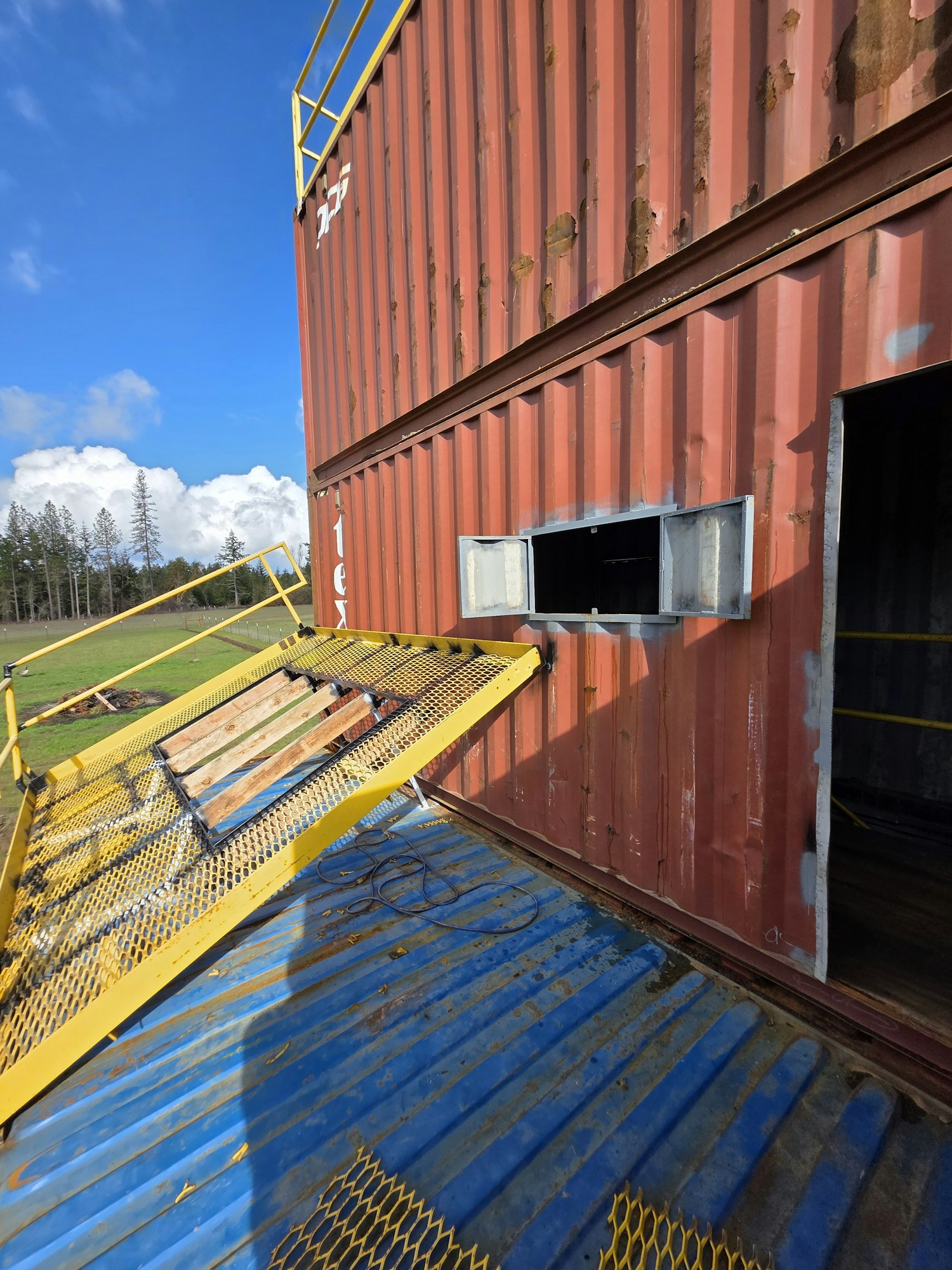 A red container with a window and a yellow metal ramp on a blue surface under a clear blue sky.