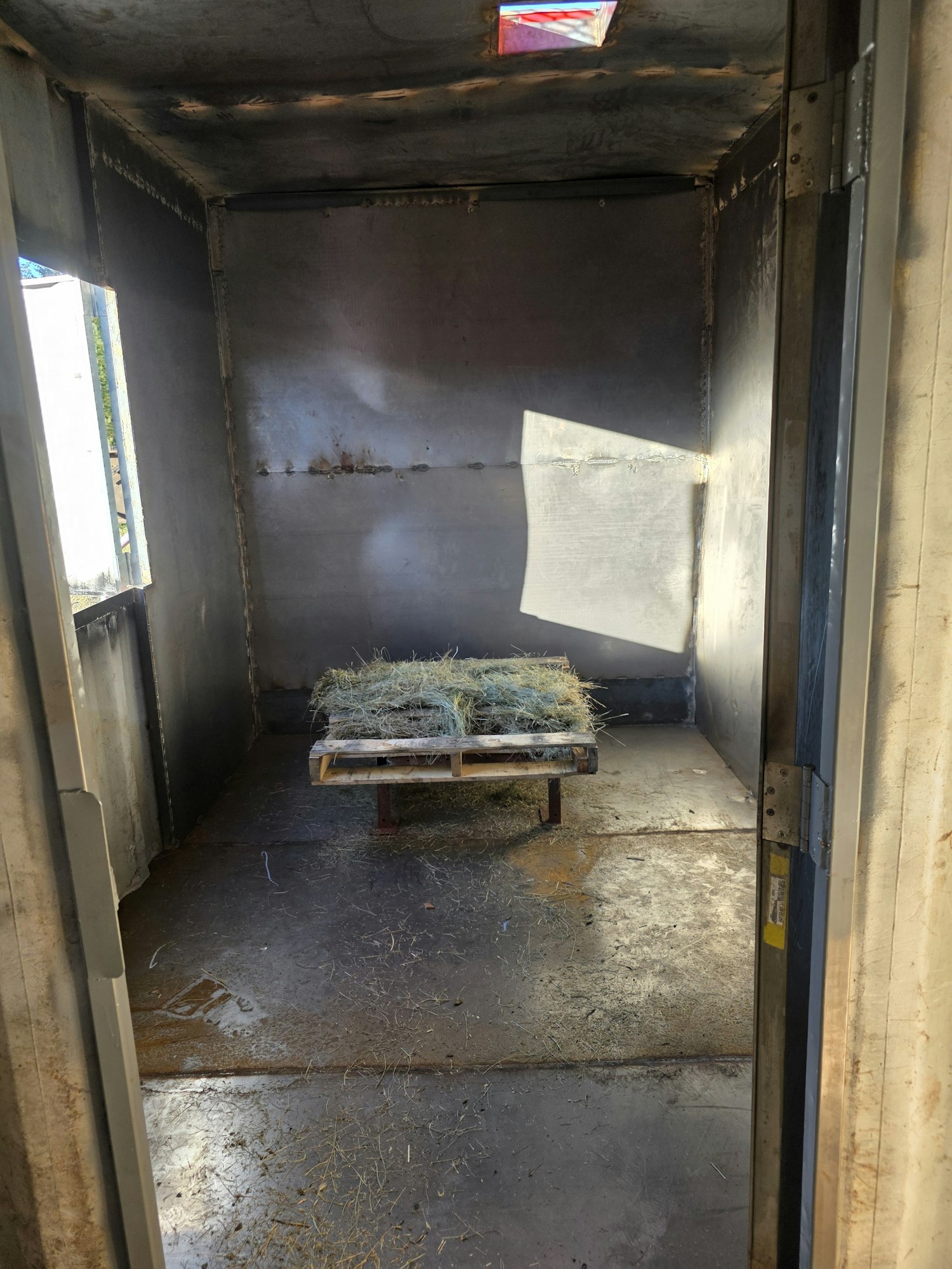 An empty metal container with hay bales on a wooden pallet inside, lit by sunlight.
