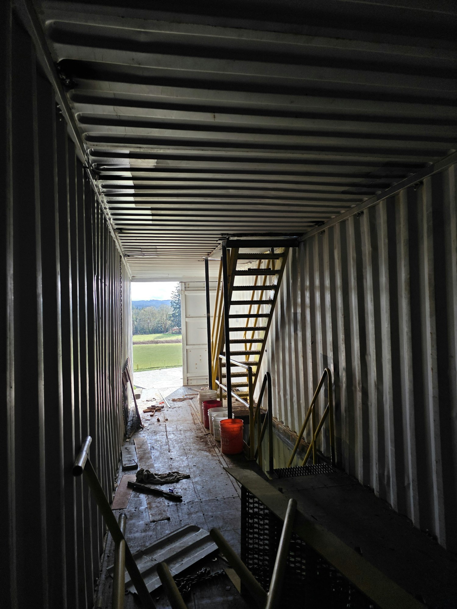 Interior of a shipping container with a ladder, scattered tools, and an open door showing grass outside.