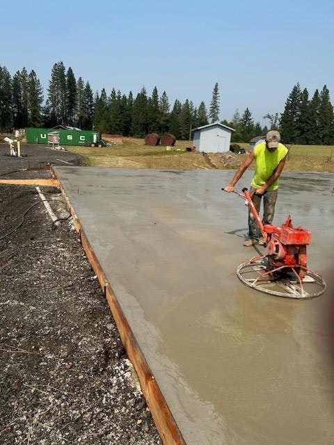 A person smooths wet concrete with a power trowel on a construction site, surrounded by trees and containers.