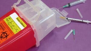 Red sharps container with biohazard label, surrounded by used syringes and needles on a purple background.