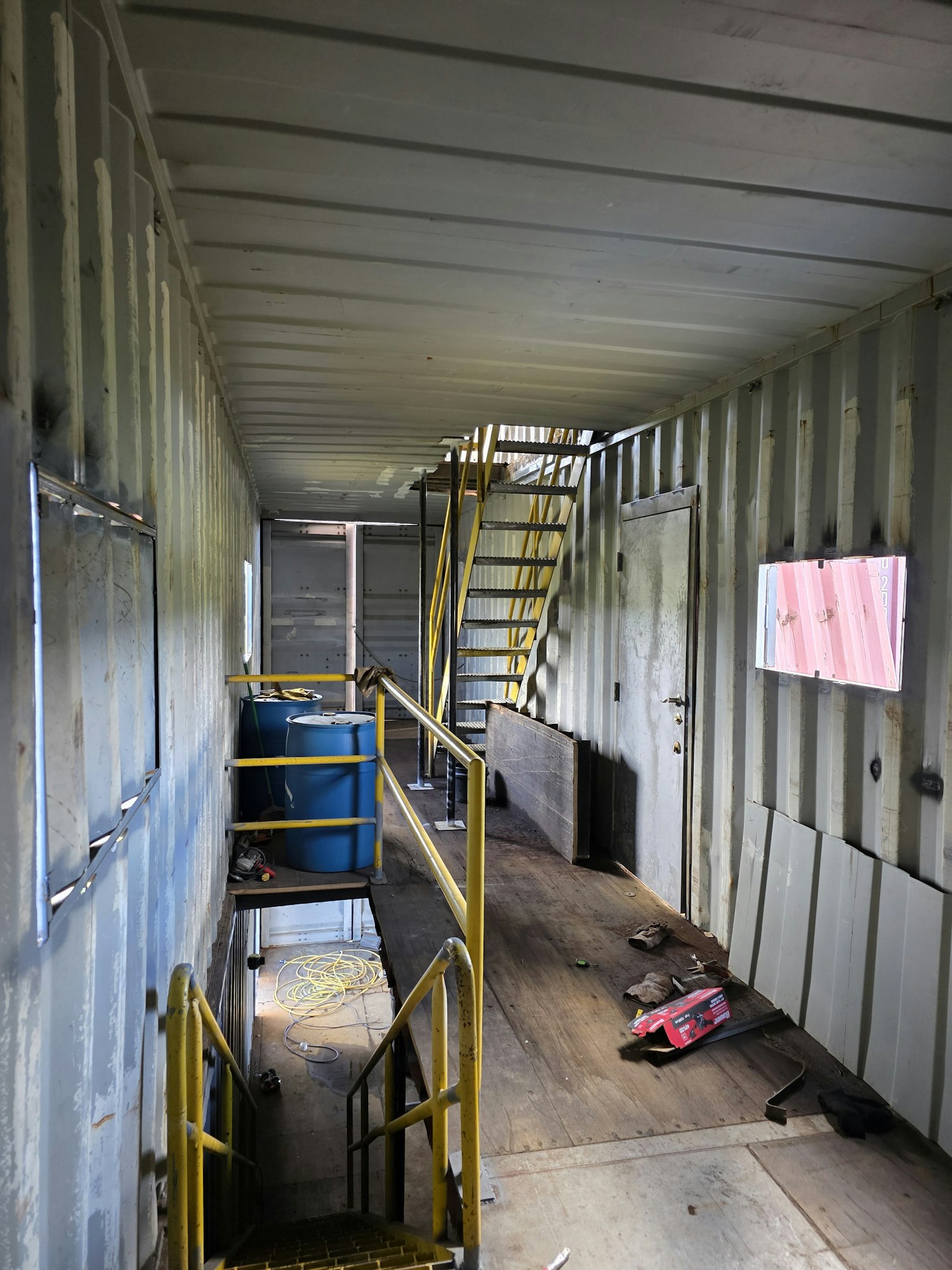 Interior of a metal container with a staircase, yellow railings, blue barrel, and scattered tools.