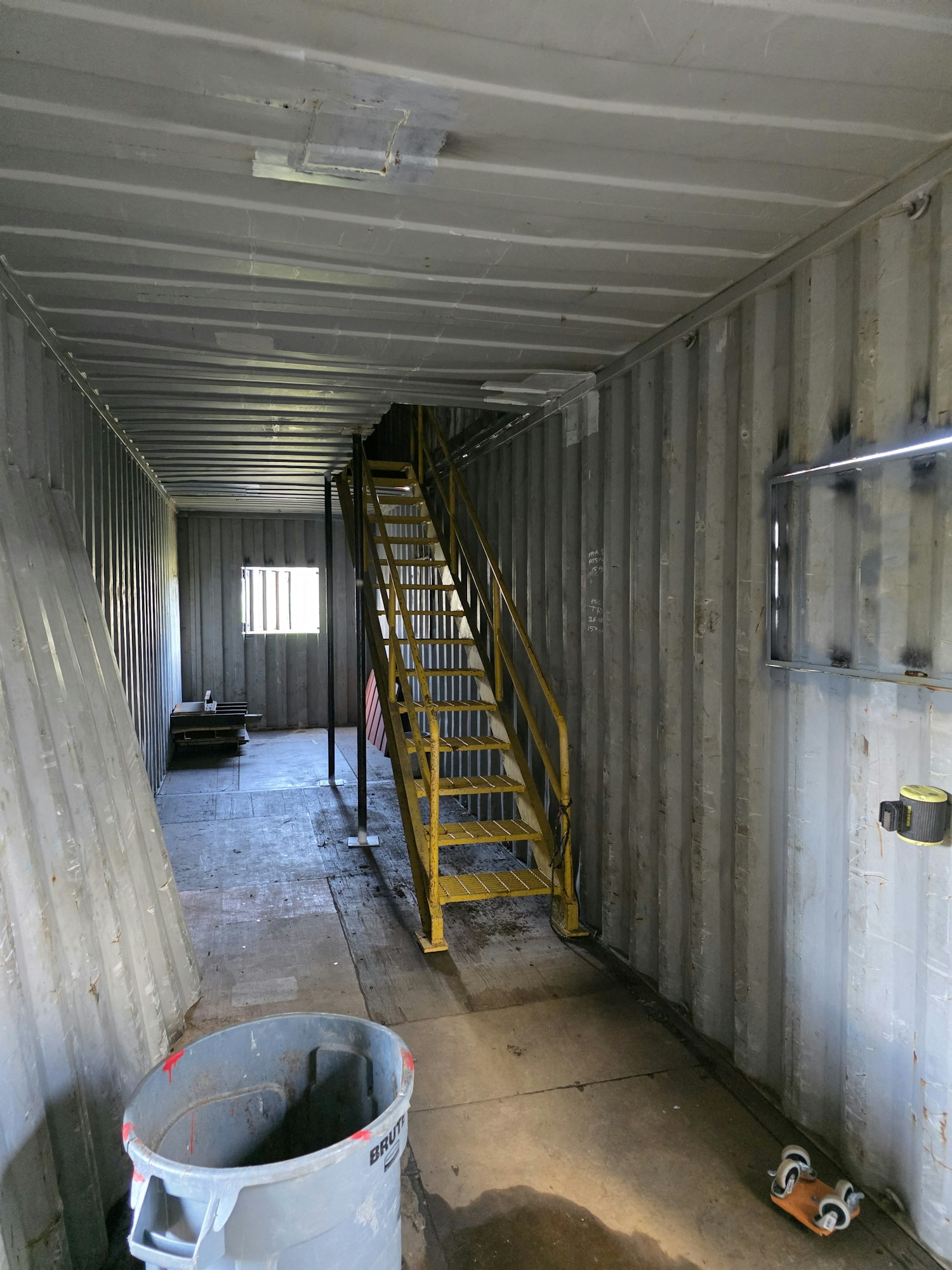 Industrial space with a yellow metal staircase, a trash bin, and metal walls, viewed sideways.