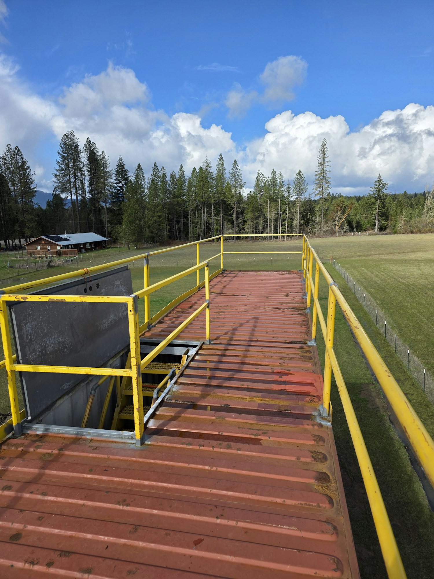 A metal structure with yellow railings, beside a grassy field and tree line under a partly cloudy sky.