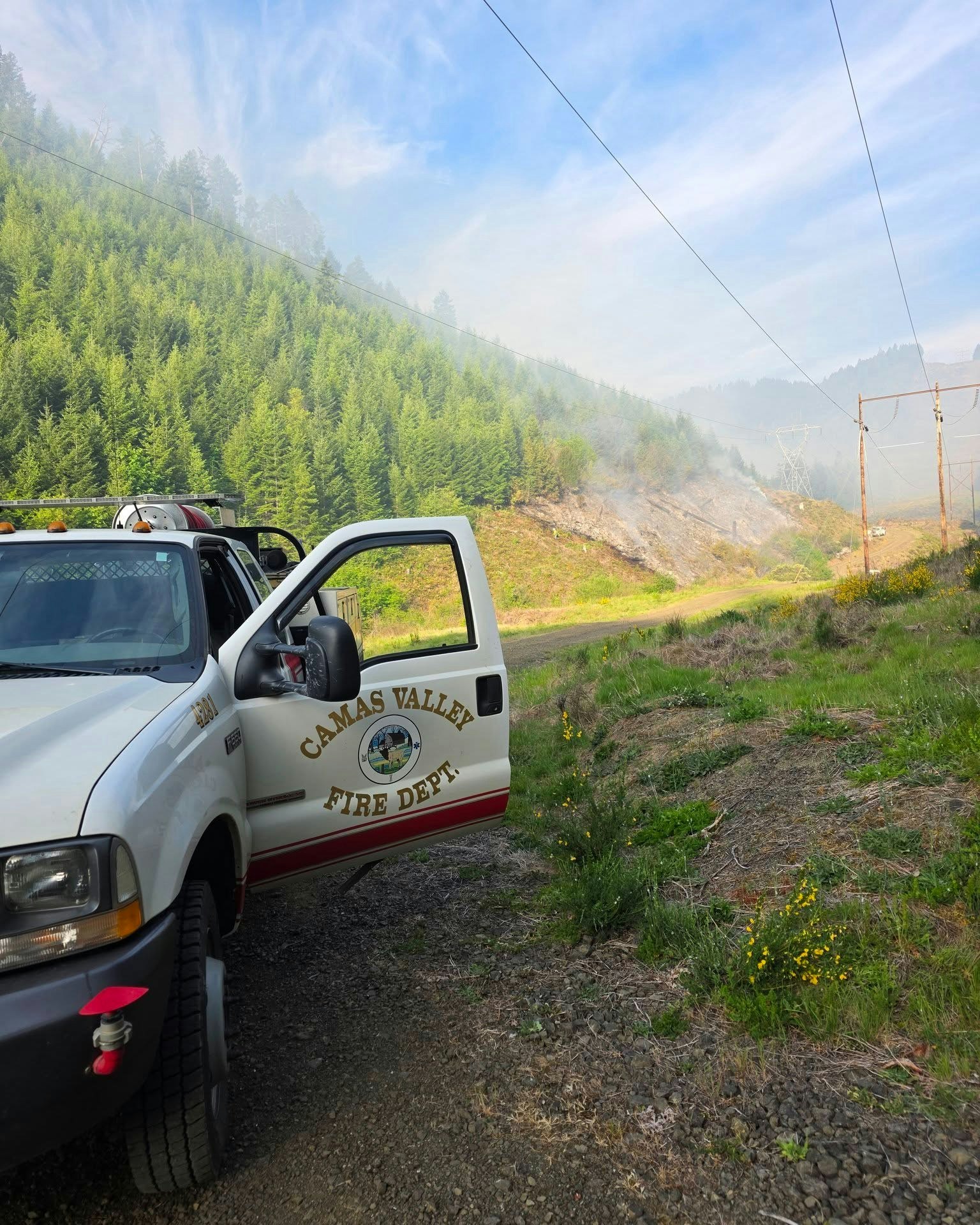 A fire department truck is parked near a hillside with smoke and overhead power lines.