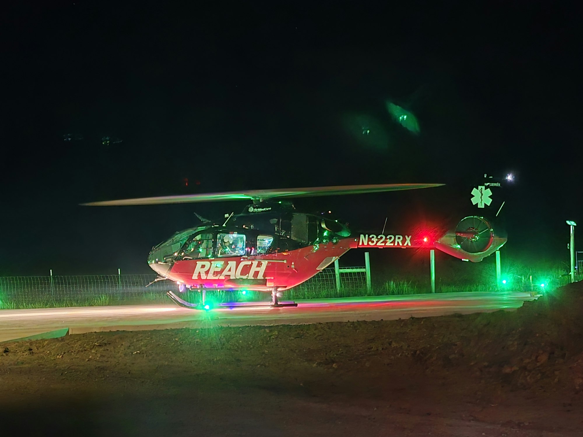 A red and black helicopter with "REACH" on the side, lit by green lights, on a helipad at night.